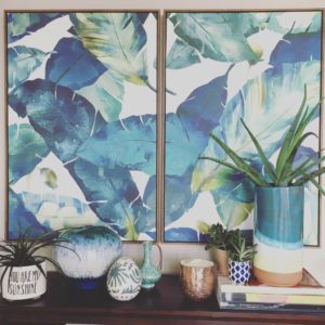 Behind a shelf of potted succulents and sculptural objects, two large framed canvases hang side by side and depict oversized, tropical foliage.