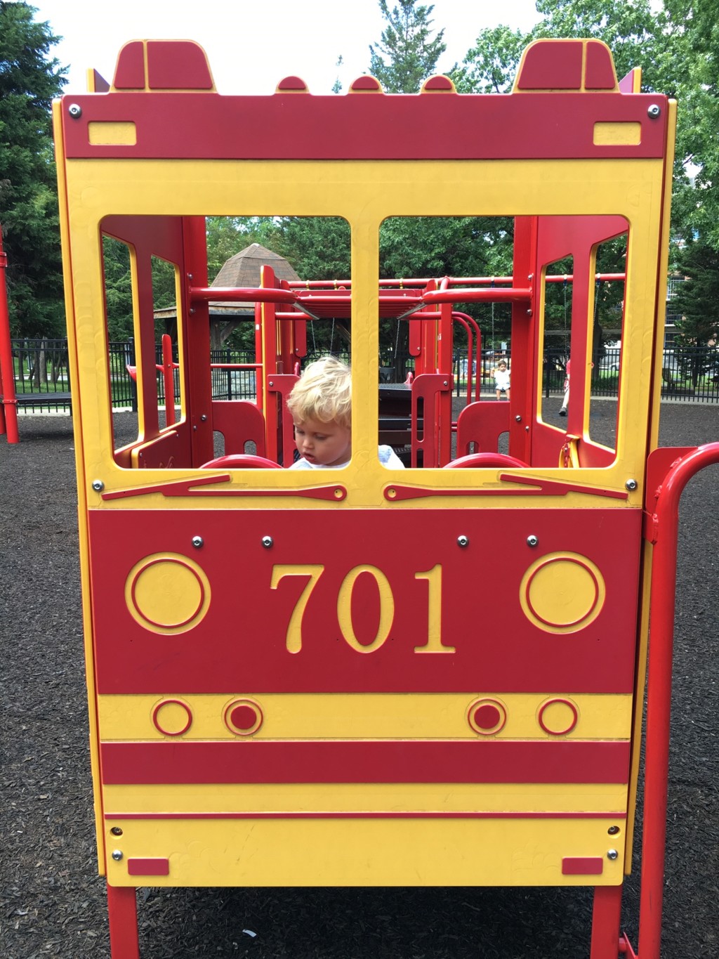 At a playground, a toddler sits behind the steering wheel of a large bright red and yellow fire engine.
