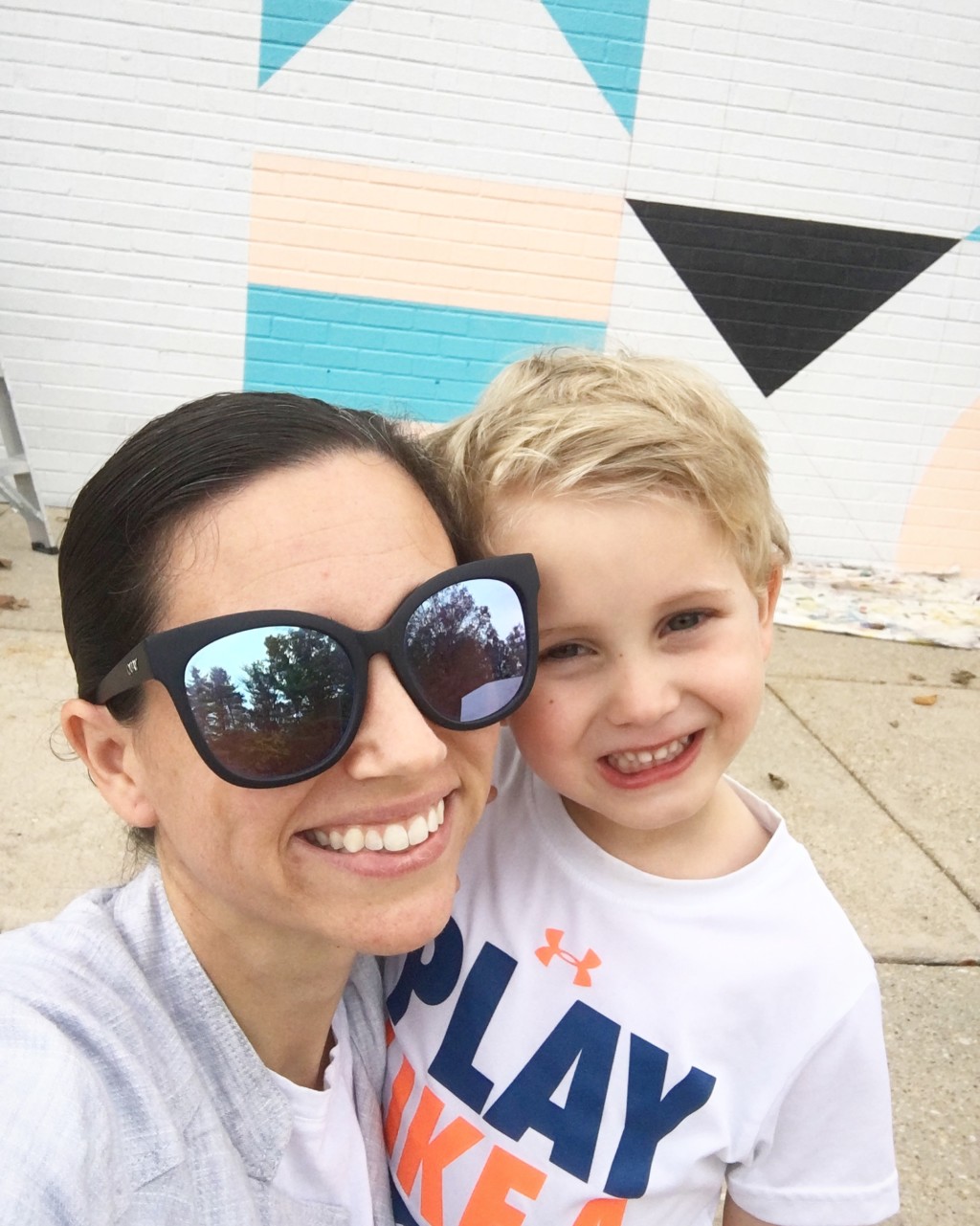 A mother and young son pose together on a sidewalk outside for a happy and smiling selfie.