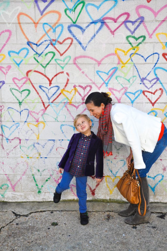 A smiling mother bends down towards her young son, while standing in front of a mural of multicolored hearts.