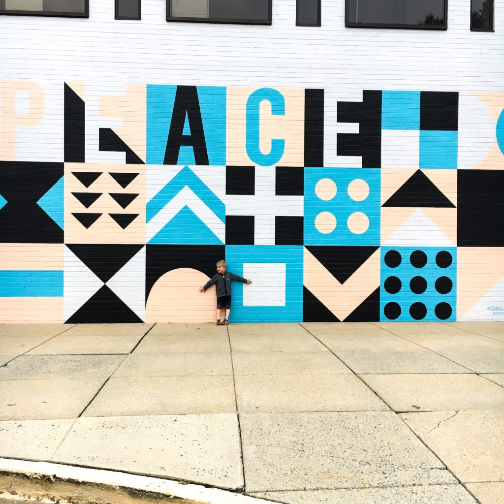 A young boy stands with his back against a large scale mural that reads P-E-A-C-E.