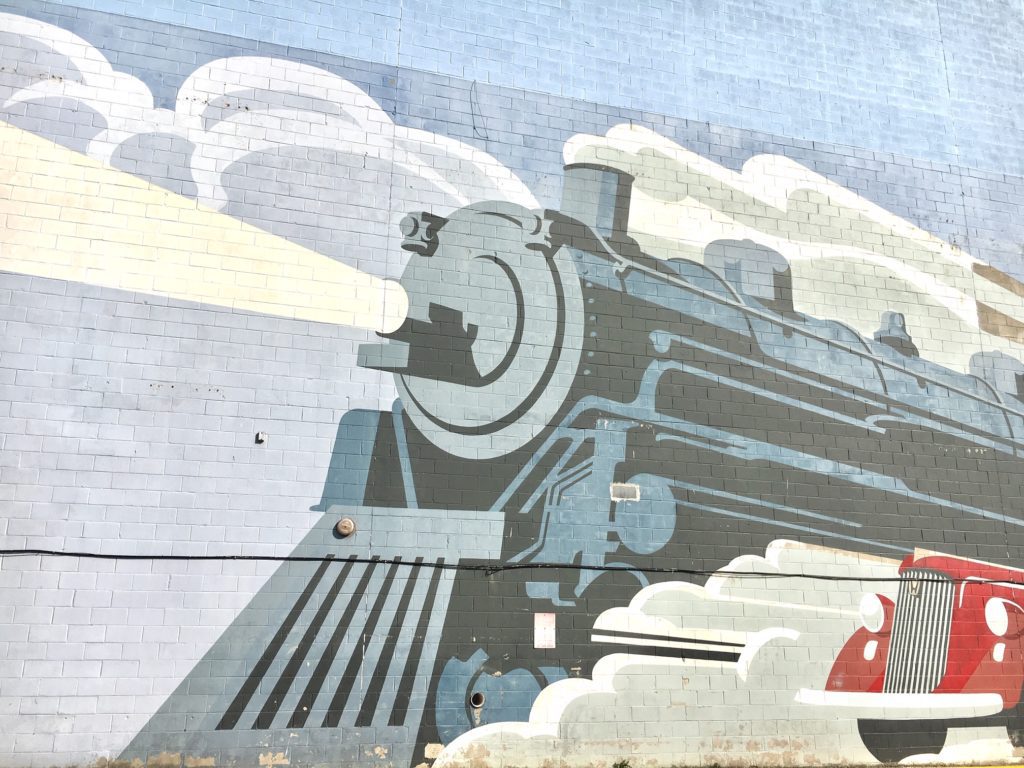 A large scale mural of a steam engine train and a red classic car are painted on the side of a building's brick wall.