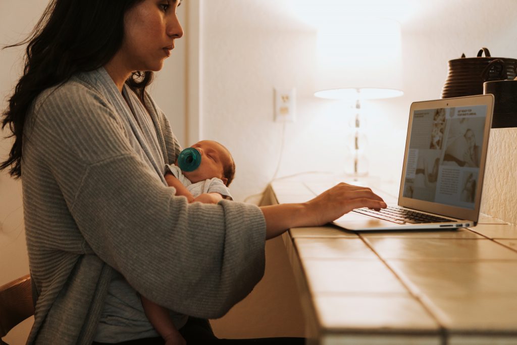 A woman on maternity leave does work on a laptop while holding her infant.