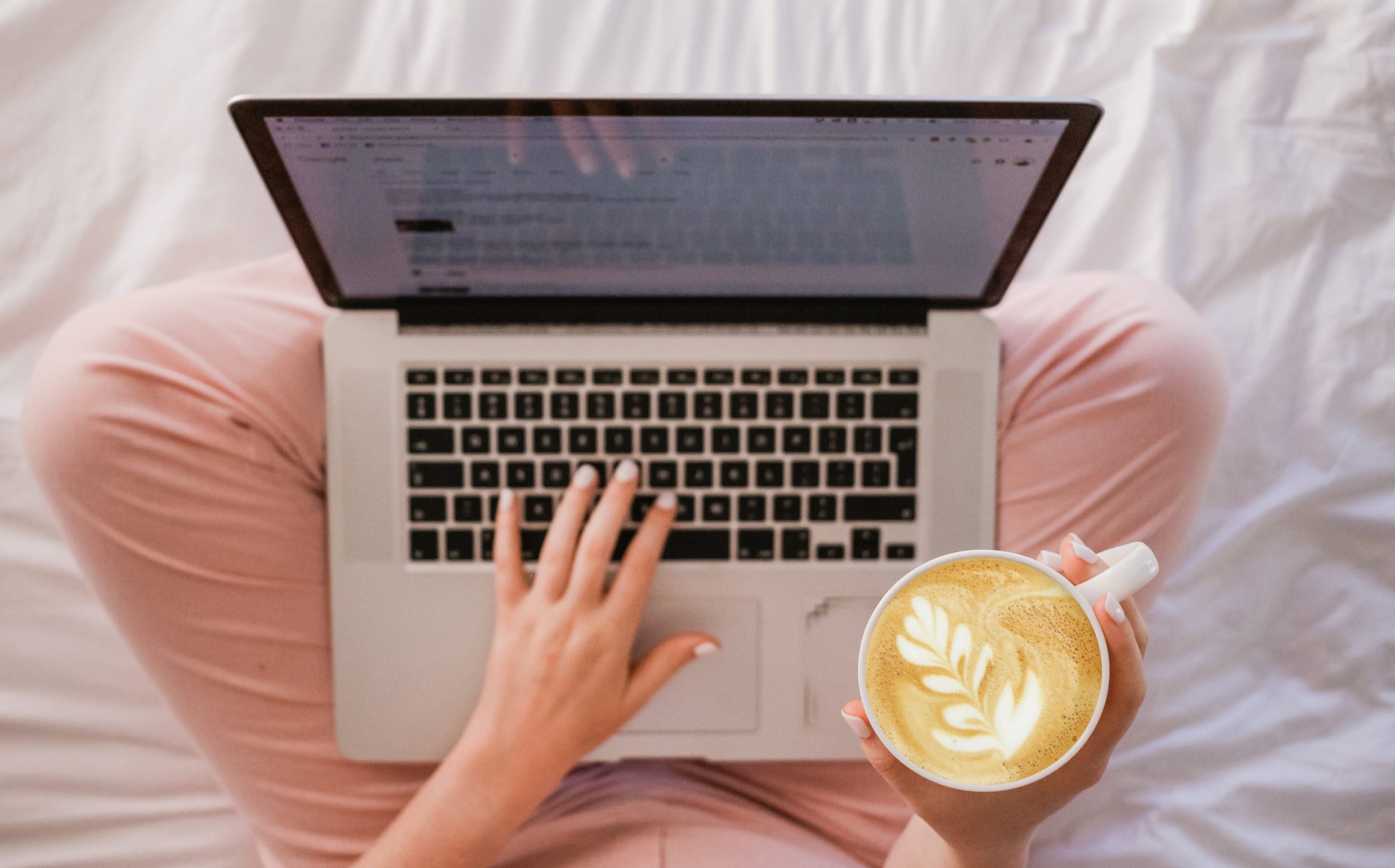 a woman holds a latte and laptop in bed