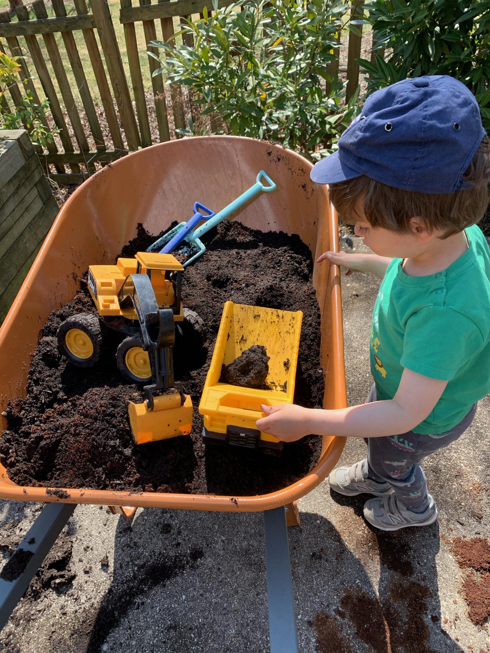 A young boy looks at his toys in a wheelbarrow full of dirt.