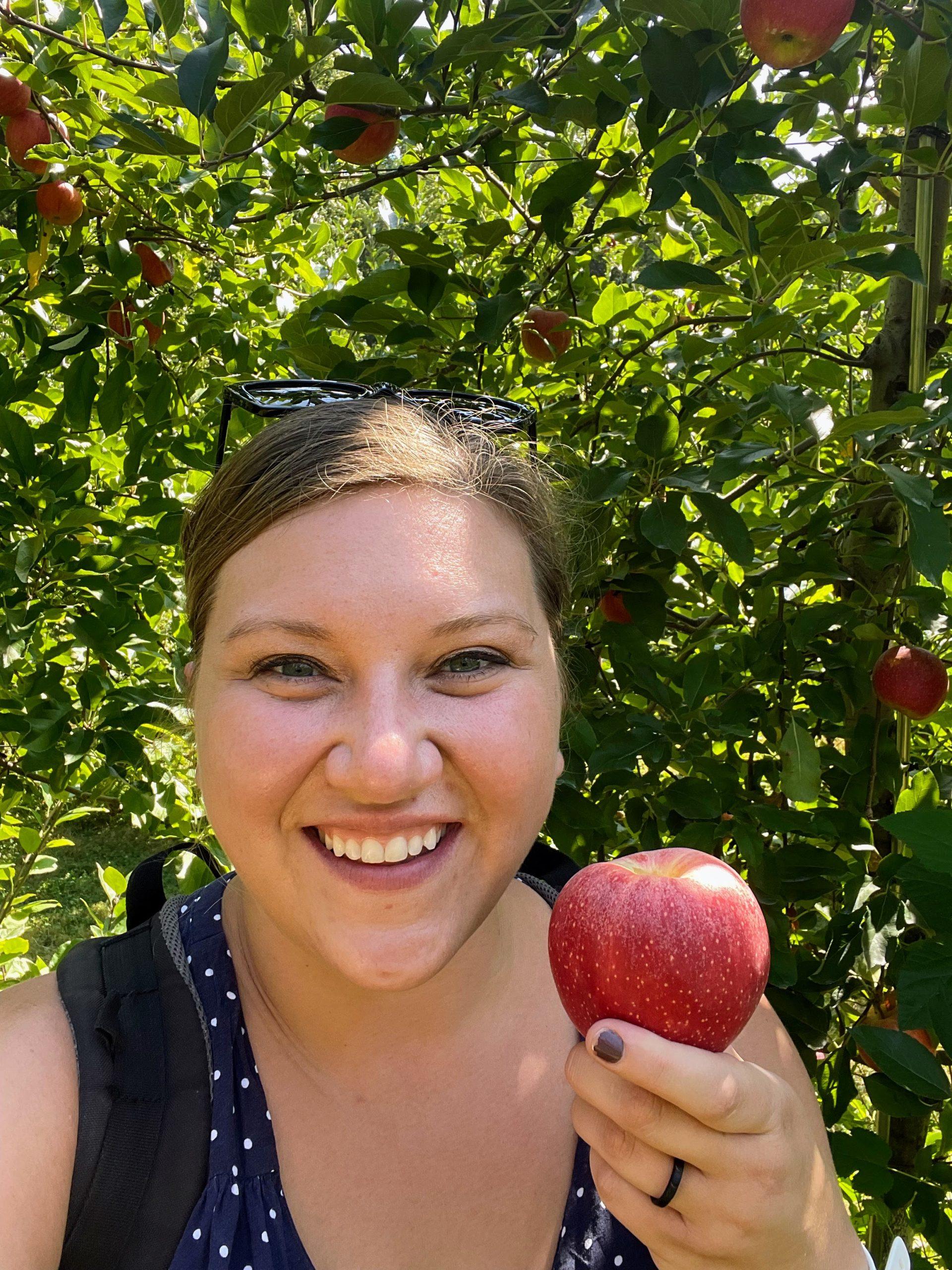 A woman is smiling and standing in an apple orchard holds one red apple in her hand