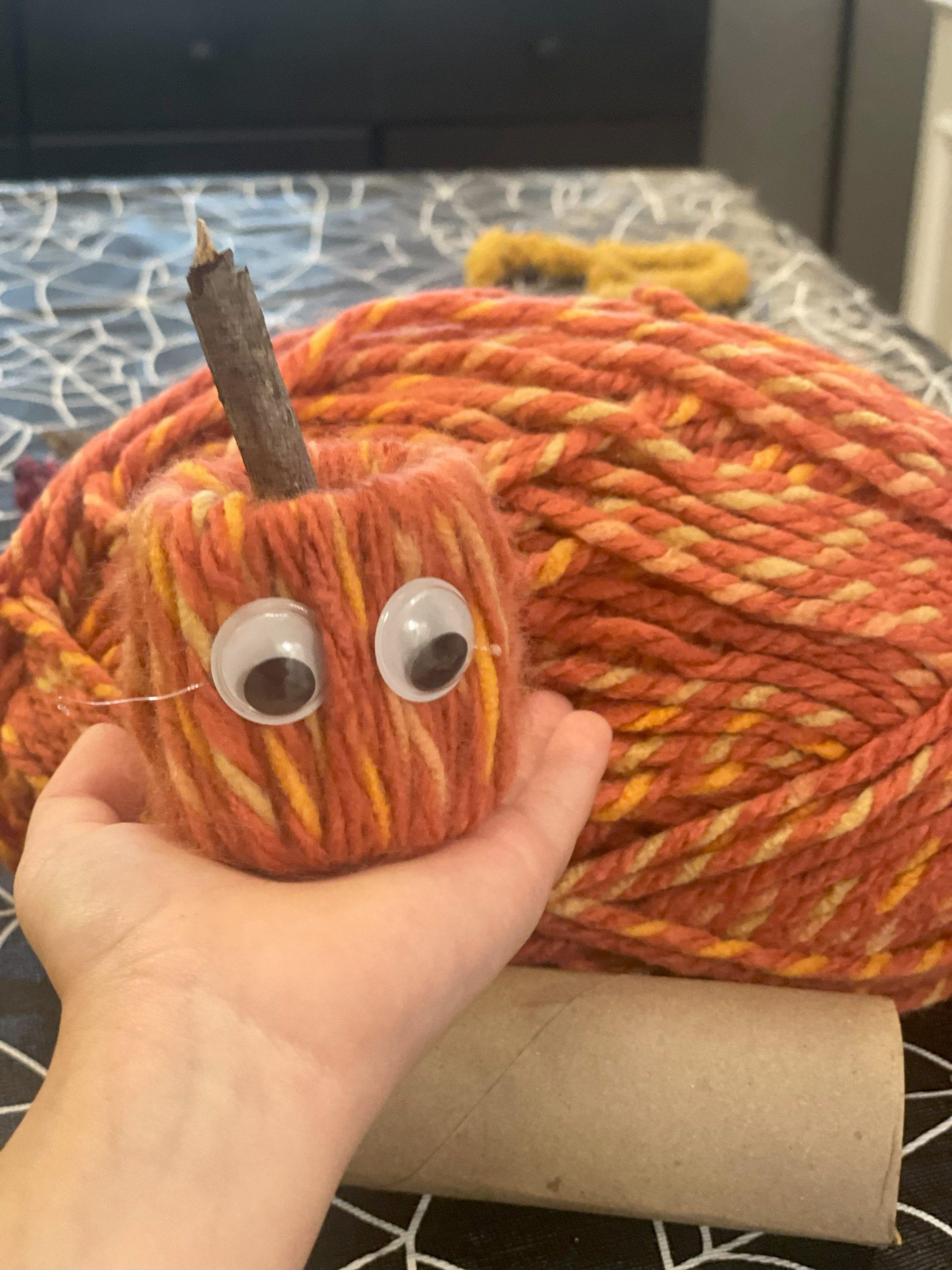 A child is holding an orange yarn pumpkin with googly eyes in their hand. 